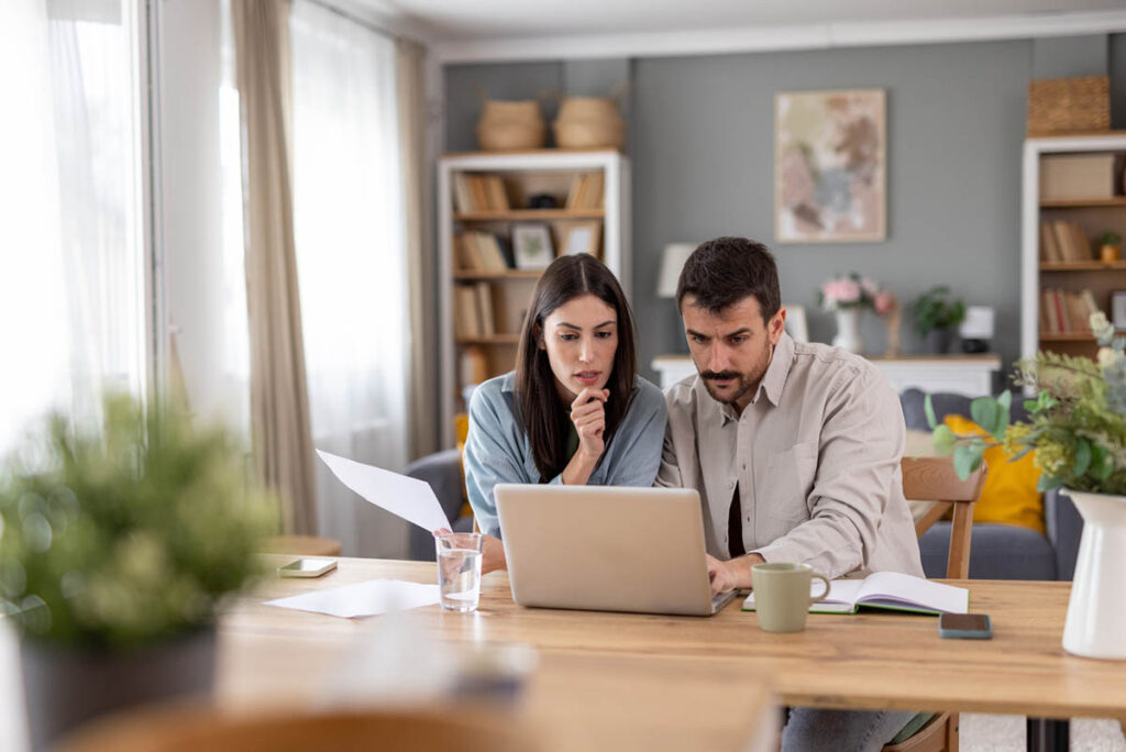 A couple is deep in thought while reviewing documents on a laptop at home.