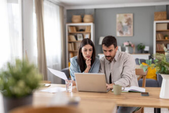 A couple is deep in thought while reviewing documents on a laptop at home.