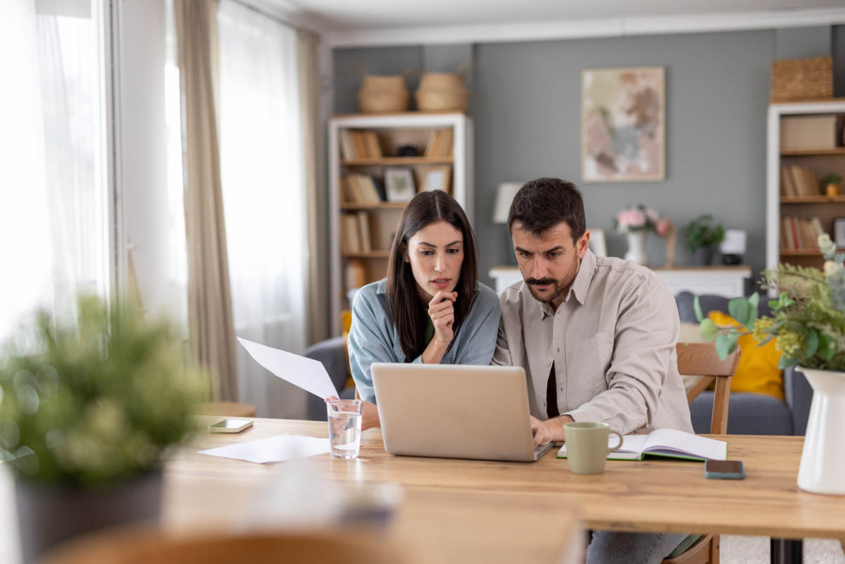 A couple is deep in thought while reviewing documents on a laptop at home.